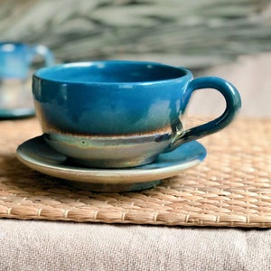 May include: A handmade ceramic teacup and saucer set. The cup and saucer are a vibrant teal color with a brown and gray rim. The cup has a rounded shape with a curved handle. The set is placed on a woven mat.