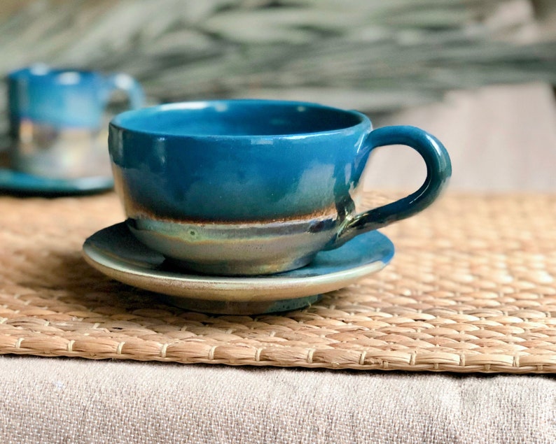 May include: A handmade ceramic teacup and saucer set. The cup and saucer are a vibrant teal color with a brown and gray rim. The cup has a rounded shape with a curved handle. The set is placed on a woven mat.