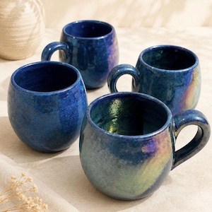 May include: Four blue ceramic mugs with rounded bodies and curved handles. The mugs have a glossy, iridescent finish that reflects shades of green and purple. The mugs are arranged on a neutral surface.