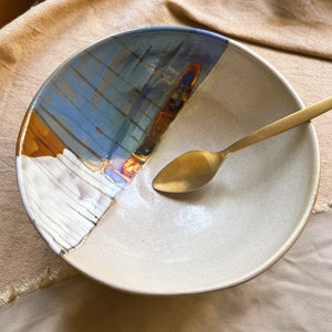 May include: A white ceramic bowl with a blue, orange, and white glaze design. A gold spoon is resting inside the bowl.
