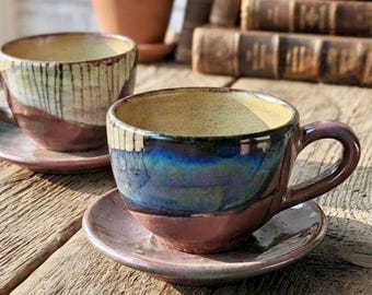 May include: Two ceramic tea cups with matching saucers, each featuring a glossy glaze that transitions from a deep blue and black upper section to a warm, earthy reddish-brown base, with a light cream-coloured interior and a sturdy side handle.
