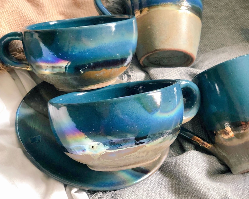May include: Handmade ceramic teacups and saucers in shades of teal and blue with iridescent accents. The cups have a rounded shape with a handle, and the saucers are circular. The glaze creates a unique, artistic look. A set of handcrafted pottery.