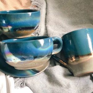 May include: A collection of handmade ceramic mugs and a saucer. The mugs are a vibrant teal color with iridescent and metallic accents. The set includes a mug with a handle and a matching saucer, perfect for enjoying coffee or tea.
