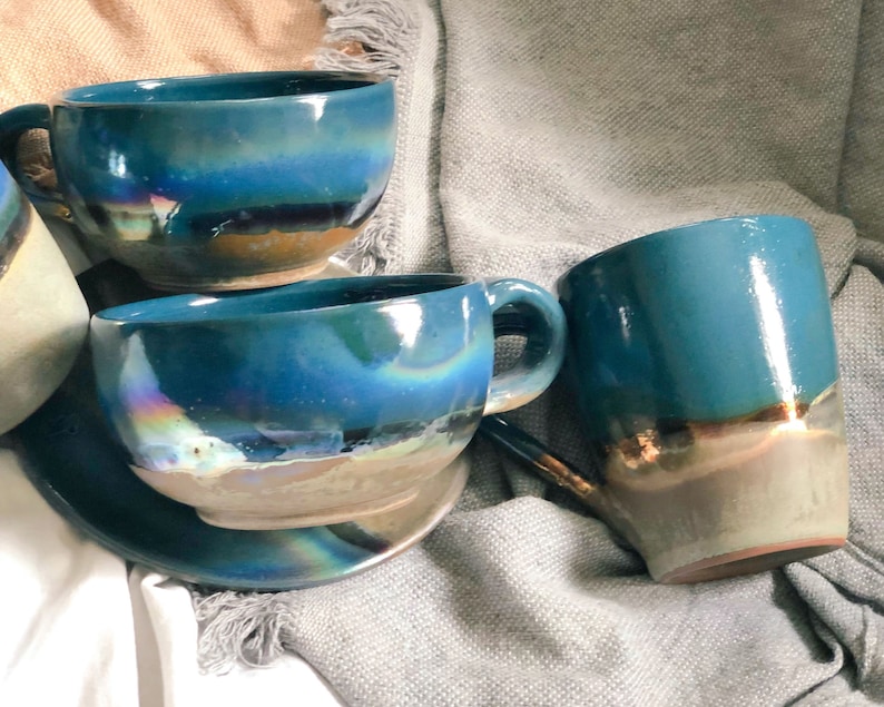 May include: A collection of handmade ceramic mugs and a saucer. The mugs are a vibrant teal color with iridescent and metallic accents. The set includes a mug with a handle and a matching saucer, perfect for enjoying coffee or tea.