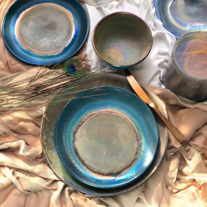 May include: A collection of iridescent ceramic tableware, including plates and a bowl, in shades of teal, blue, and green. The plates have a layered design with a metallic sheen. A wooden knife and peacock feather add to the aesthetic.