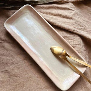Könnte beinhalten: Ein rechteckiges Serviertablett aus Keramik mit hellrosa Glasur. Das Tablett hat einen dezenten irisierenden Glanz und liegt auf einer beigefarbenen Stoffoberfläche. Zwei goldene Löffel liegen auf dem Tablett.