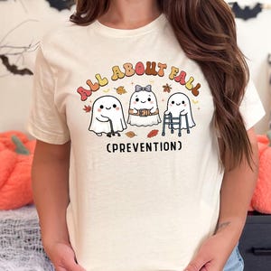 May include: A cream-colored short-sleeve t-shirt featuring a graphic of three ghosts with the text "ALL ABOUT FALL (PREVENTION)". The ghosts are depicted with a cane, a belt, and a walker, surrounded by small autumn leaves in orange and brown.