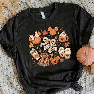 May include: A dark charcoal gray t-shirt featuring a graphic print with the text 'TIS THE SEASON' surrounded by autumn-themed illustrations including pumpkins with mouse ears, a plaid scarf, slippers, a coffee cup, and fall leaves.