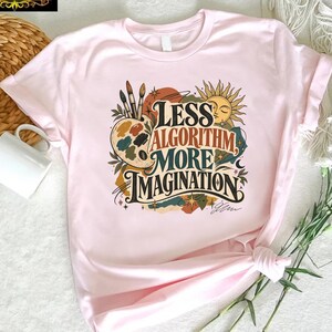 May include: A light pink short-sleeve t-shirt featuring a graphic print with the text "LESS ALGORITHM MORE IMAGINATION" in a stylized font. The design includes illustrations of a paint palette, brushes, a sun, and various decorative elements.