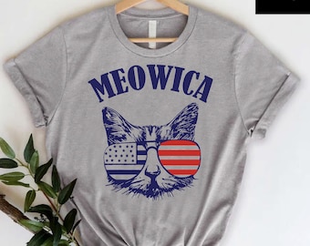 May include: A heather gray t-shirt featuring a cat face wearing sunglasses with the American flag design. The word "MEOWICA" is printed above the cat's face in navy blue. The shirt is hanging on a wooden hanger.