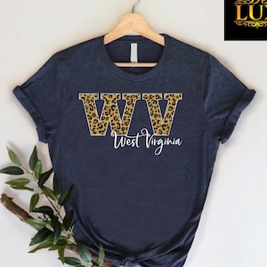 May include: Navy blue t-shirt with the letters "WW" in a leopard print design, with the words "West Virginia" in white script below. The shirt is displayed on a wooden hanger.