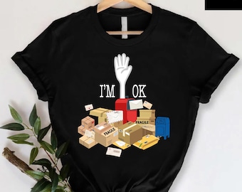 May include: Black t-shirt with the text "I'M OK" and a white hand graphic. The graphic includes a pile of packages, envelopes, and a mailbox. The shirt is hanging on a wooden hanger.