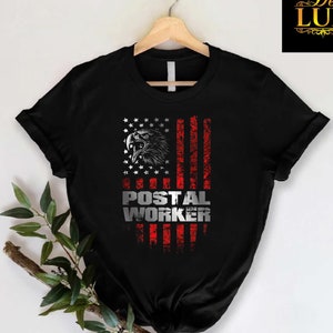 May include: Black t-shirt featuring an American flag design with a distressed look. The flag incorporates a detailed eagle head and the text "POSTAL WORKER" in a silver, bold font. The shirt is displayed on a wooden hanger.
