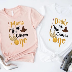 May include: Two short-sleeved t-shirts, one pink and one white, with a Harry Potter theme. The pink shirt says "Mama Of The Chosen One" and the white shirt says "Daddy Of The Chosen One". Each shirt features a golden snitch and a wizard's hat.