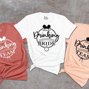 May include: Three short-sleeved t-shirts in coral, white, and peach. Each shirt features a black Mickey Mouse silhouette and the words "Drinking Team" or "Drinking Bride" in a circular design. The shirts are knotted at the bottom.