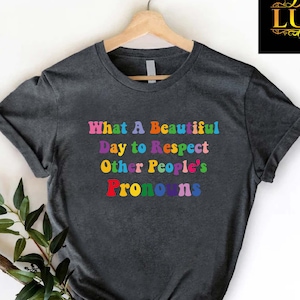 May include: Dark gray t-shirt with the text "What A Beautiful Day to Respect Other People's Pronouns" in rainbow colors. The shirt is hanging on a wooden hanger against a white background. A logo is in the upper right corner.