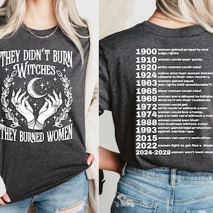 May include: Dark gray t-shirt with the text "THEY DIDN'T BURN WITCHES THEY BURNED WOMEN" in white. The design includes a crescent moon, stars, and hands. The back lists women's rights milestones from 1900 to 2028.