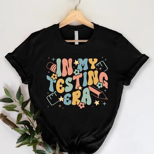 May include: Black t-shirt with the text "IN MY TESTING ERA" in a retro font. The design includes colorful illustrations of a pencil, ruler, and flowers. The shirt is hanging on a wooden hanger.