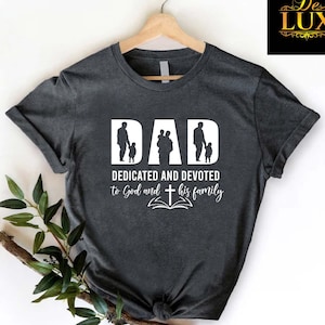 May include: Dark gray t-shirt with the word "DAD" in white block letters, featuring silhouettes of a father and children. Below, the text reads "DEDICATED AND DEVOTED to God and his family" with a cross and open book graphic.