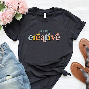 May include: Black t-shirt with the text "let's get creative" in colorful, retro-style lettering. The shirt is paired with blue jeans, sandals, and pink flowers, creating a casual, artistic aesthetic.