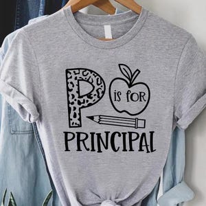 May include: A gray t-shirt with the words "P is for Principal" in black. The letter "P" has a leopard print design. An apple and pencil graphic are also included in the design. The shirt is hanging on a wooden hanger.