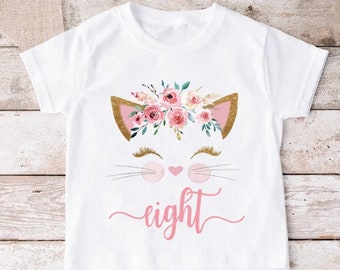 May include: White t-shirt featuring a cat face design with gold ears, pink nose, and floral crown. The word "eight" is written in pink script below the cat face. A cute and whimsical design for a child's birthday.