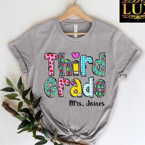 May include: A gray t-shirt with the words "Third Grade" in colorful, patterned letters and a pink heart. The text "Mrs. Jones" is below the main text. The shirt is hanging on a wooden hanger.