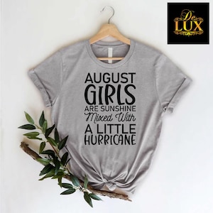 May include: A heather gray t-shirt with the text "AUGUST GIRLS ARE SUNSHINE Mixed With A LITTLE HURRICANE" in black. The shirt is hanging on a wooden hanger with a sprig of greenery.