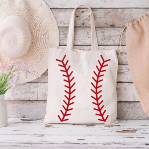 May include: A cream-colored tote bag with red baseball stitching design. The bag has long handles and is made of a sturdy fabric. The design is a bold, graphic representation of a baseball.