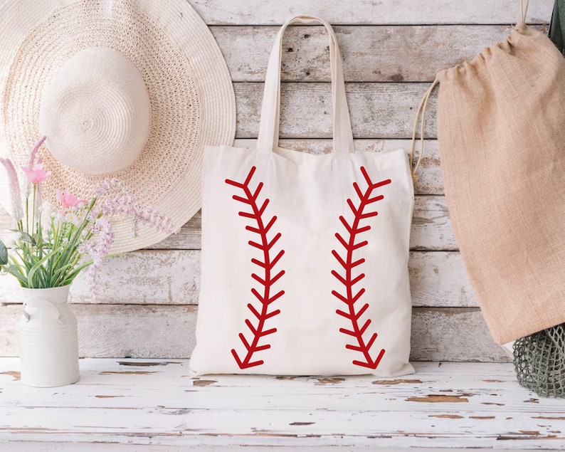 May include: A cream-colored tote bag with red baseball stitching design. The bag has long handles and is made of a sturdy fabric. The design is a bold, graphic representation of a baseball.
