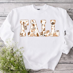 May include: White sweatshirt with the word "FALL" in large, block letters. Each letter is filled with a fall-themed pattern of leaves and acorns in shades of brown and beige. The sweatshirt has long sleeves.