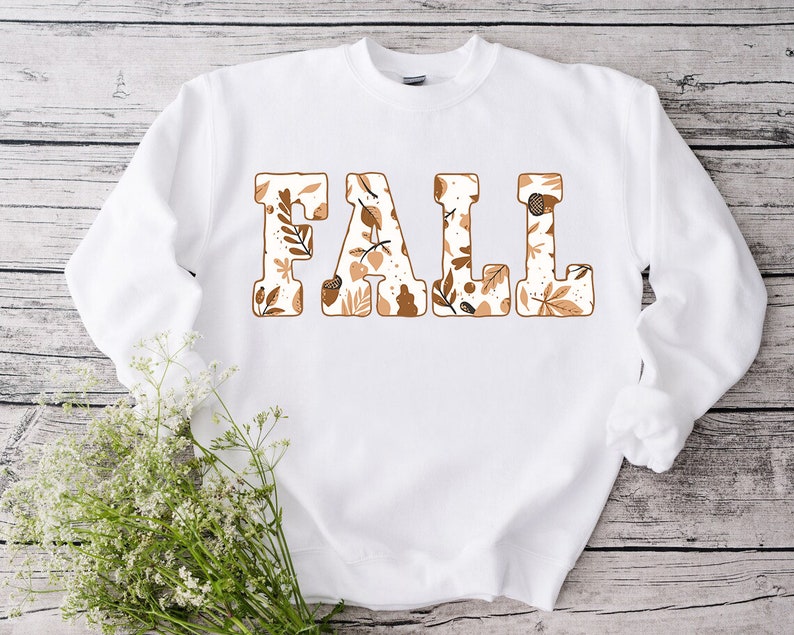 May include: White sweatshirt with the word "FALL" in large, block letters. Each letter is filled with a fall-themed pattern of leaves and acorns in shades of brown and beige. The sweatshirt has long sleeves.