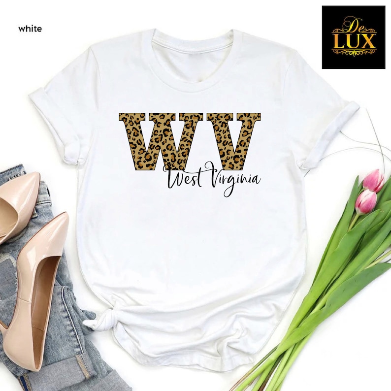 May include: White t-shirt featuring the letters "WV" in a leopard print design, with "West Virginia" written below in a script font. The shirt is displayed with a pair of beige heels and a sprig of pink flowers.