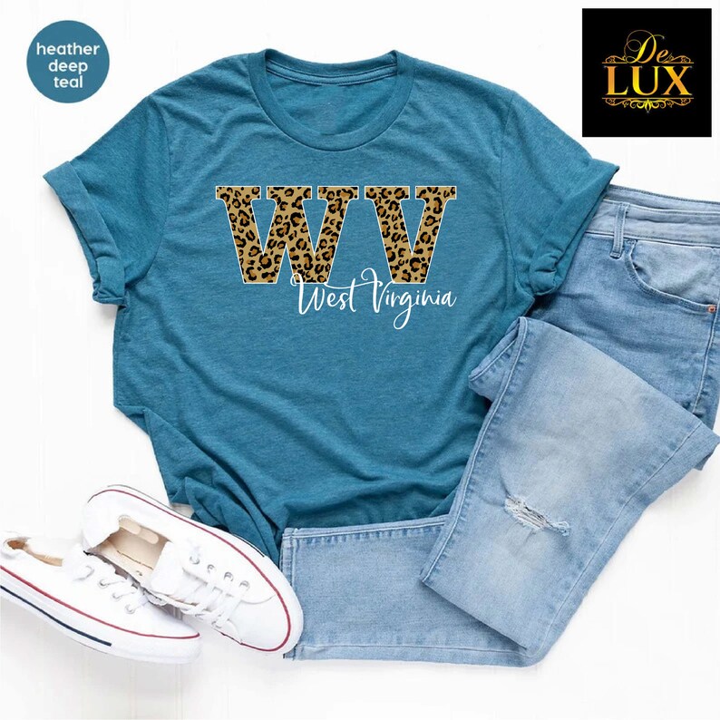May include: A teal t-shirt with the letters "WV" in a leopard print design, with the words "West Virginia" in a script font. The shirt is paired with blue jeans and white sneakers.