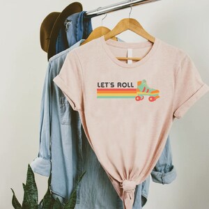 May include: A light pink t-shirt with the text "LET'S ROLL" above a retro roller skate graphic. The graphic features a teal skate with orange wheels and a rainbow stripe design. The shirt is hanging on a wooden hanger.
