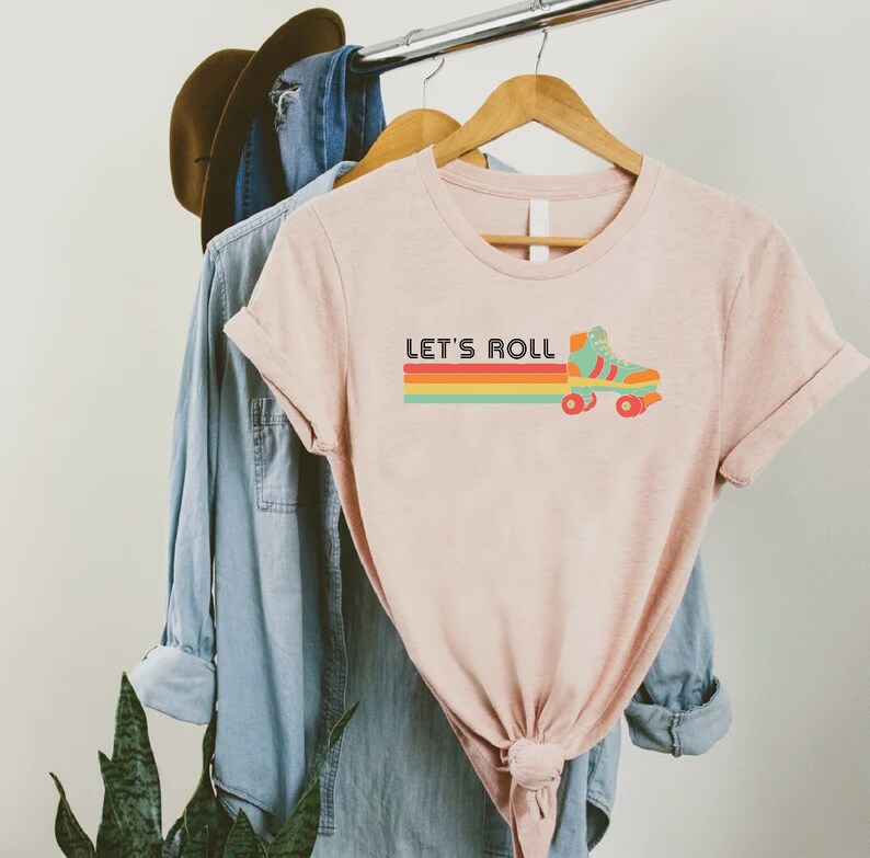 May include: A light pink t-shirt with the text "LET'S ROLL" above a retro roller skate graphic. The graphic features a teal skate with orange wheels and a rainbow stripe design. The shirt is hanging on a wooden hanger.