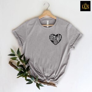 May include: A heather gray t-shirt with a black heart-shaped graphic that reads "LATINA". The shirt is displayed on a wooden hanger against a white background, with a sprig of greenery in the foreground.