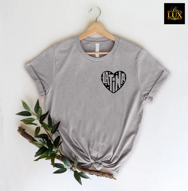 May include: A heather gray t-shirt with a black heart-shaped graphic that reads "LATINA". The shirt is displayed on a wooden hanger against a white background, with a sprig of greenery in the foreground.