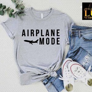 Puede incluir: Una camiseta gris claro con las palabras "AIRPLANE MODE" y la silueta de un avi&oacute;n. La camiseta se combina con vaqueros azules y zapatillas blancas. Se ve una ramita de vegetaci&oacute;n.