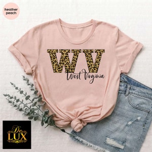 May include: A heather peach t-shirt featuring the letters "WV" in a leopard print design, with "West Virginia" written below in a script font. The shirt is paired with denim shorts.