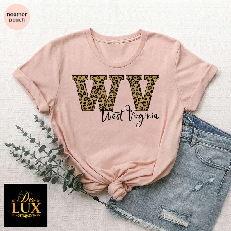 May include: A heather peach t-shirt featuring the letters "WV" in a leopard print design, with "West Virginia" written below in a script font. The shirt is paired with denim shorts.