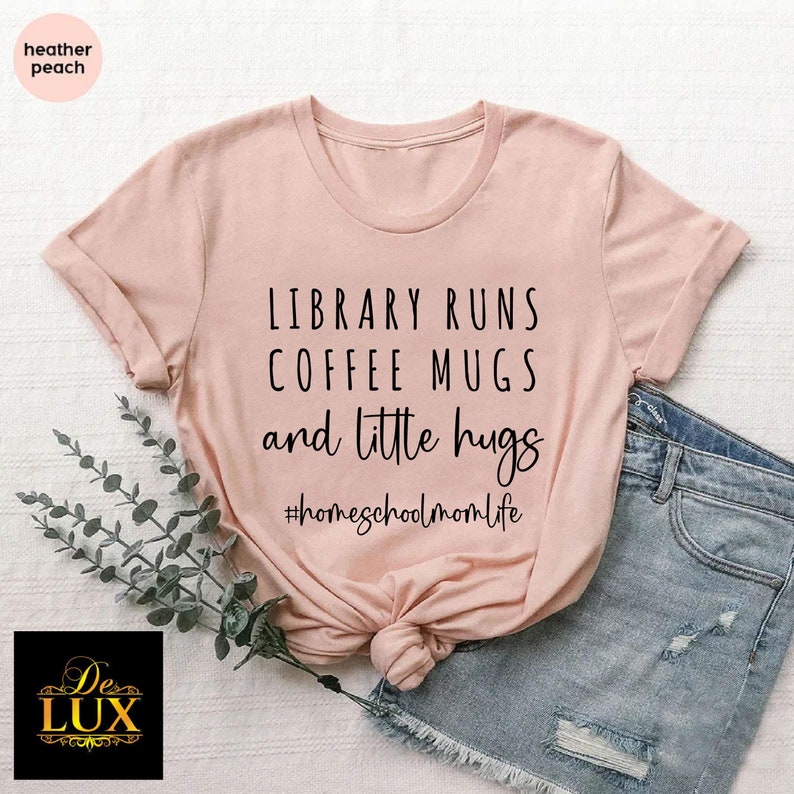 May include: Heather peach-colored t-shirt with the text "LIBRARY RUNS COFFEE MUGS and little hugs" in black. The hashtag "#homeschoolmomlife" is below the text. The shirt is knotted at the waist.