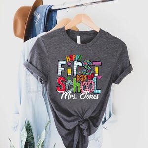 May include: A dark gray t-shirt with the words "Happy First Day of School" in colorful letters. The shirt also includes the name "Mrs. Jones" in white script. The shirt is hanging on a wooden hanger.
