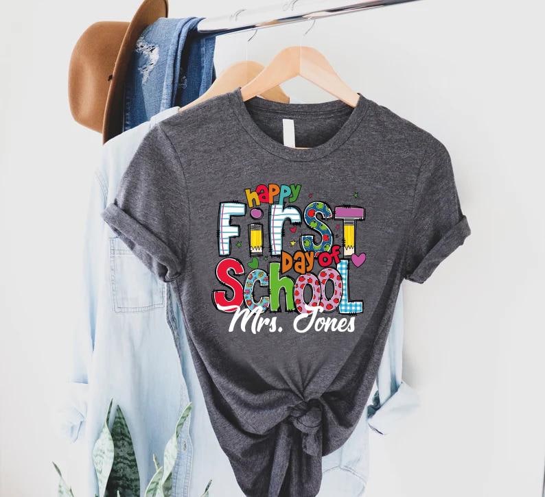 May include: A dark gray t-shirt with the words "Happy First Day of School" in colorful letters. The shirt also includes the name "Mrs. Jones" in white script. The shirt is hanging on a wooden hanger.