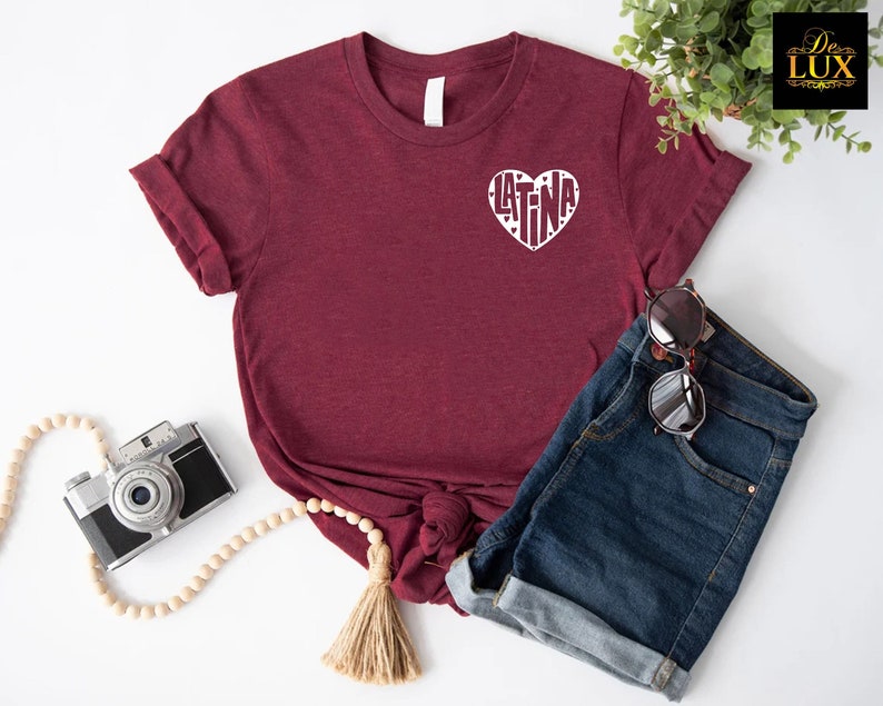 May include: Burgundy t-shirt with a white heart-shaped graphic that says "LATINA". The shirt is paired with denim shorts and sunglasses. A vintage camera and wooden beads are also in the image.