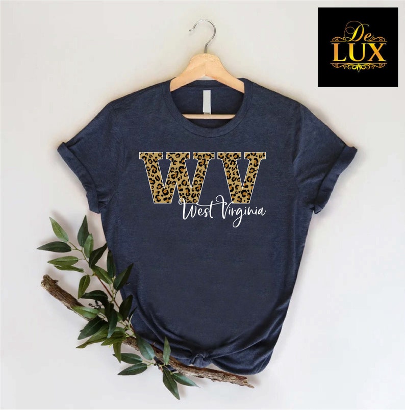 May include: Navy blue t-shirt with the letters "WW" in a leopard print design, with the words "West Virginia" in white script below. The shirt is displayed on a wooden hanger.
