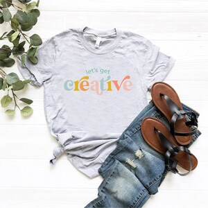 May include: A light gray t-shirt with the text "let's get creative" in colorful, retro-style lettering. The shirt is paired with blue denim jeans and black sandals with brown soles. The items are arranged on a white wooden surface.