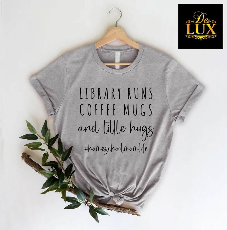 May include: A heather gray t-shirt with the text "LIBRARY RUNS COFFEE MUGS and little hugs #homeschoolmomlife". The shirt is hanging on a wooden hanger with a sprig of greenery.