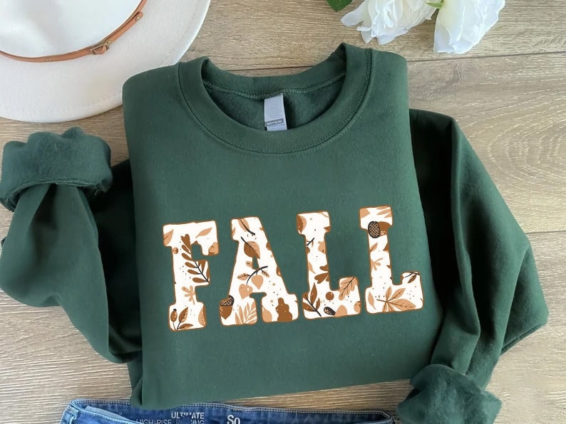May include: A dark green sweatshirt with the word "FALL" in large, block letters. The letters are filled with a pattern of fall leaves and acorns in shades of brown and cream. The sweatshirt is folded, with the sleeves and a portion of the collar visible.