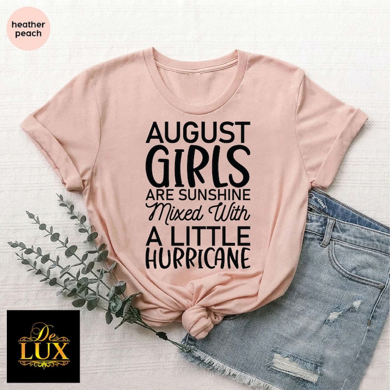 May include: A heather peach-colored t-shirt with the text "AUGUST GIRLS are sunshine mixed with a little hurricane" in black. The shirt is styled with denim shorts and a sprig of greenery.